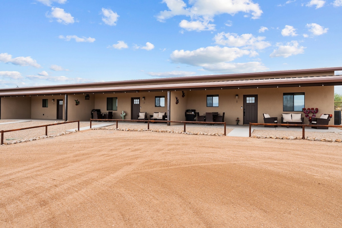 The exterior of the luxury casitas showcases a modern design with a neutral color palette. A wide, dusty pathway leads to the entrance. Seating areas with cushions and decorative accents are visible on private patios, framed by desert landscaping under a clear blue sky.