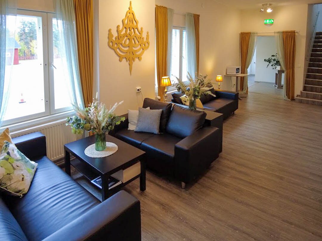 A welcoming shared lounge area features sleek black sofas arranged around a coffee table. Decorative cushions add subtle texture, while potted plants and fresh flower arrangements contribute to the inviting environment. Large windows allow natural light to fill the room, enhanced by soft drapery.