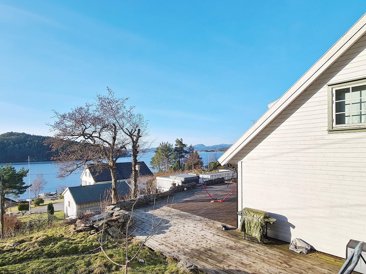 A corner view of the holiday home reveals a spacious wooden deck with seating areas overlooking the fjord. The landscape features hills and trees, with several houses dotting the shoreline in the distance against a clear blue sky.