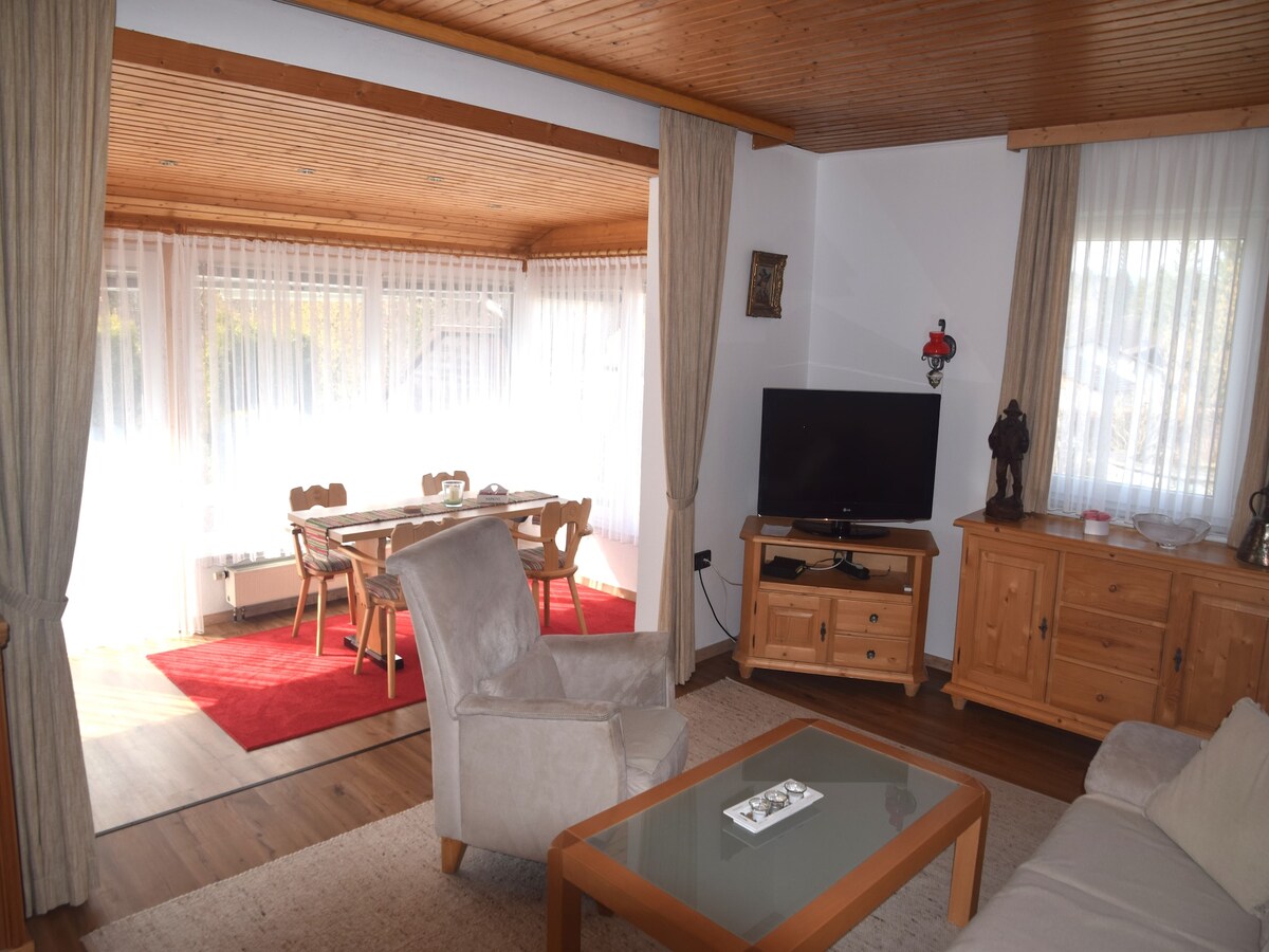 The living area features a comfortable seating arrangement with a light-colored sofa and a glass coffee table. A television is positioned against a wooden cabinet, while large windows allow ample natural light. The adjacent dining area includes a wooden table with multiple chairs, enhancing the open layout.