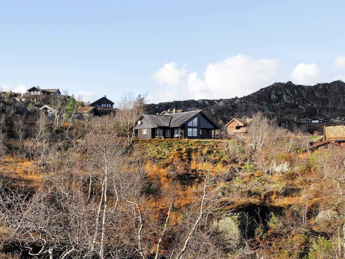 A modern holiday home with a dark wooden exterior is situated amidst a natural landscape. Surrounding greenery and rocky hills are visible, creating a rustic setting. The sky is blue with scattered clouds, enhancing the serene atmosphere of the area.