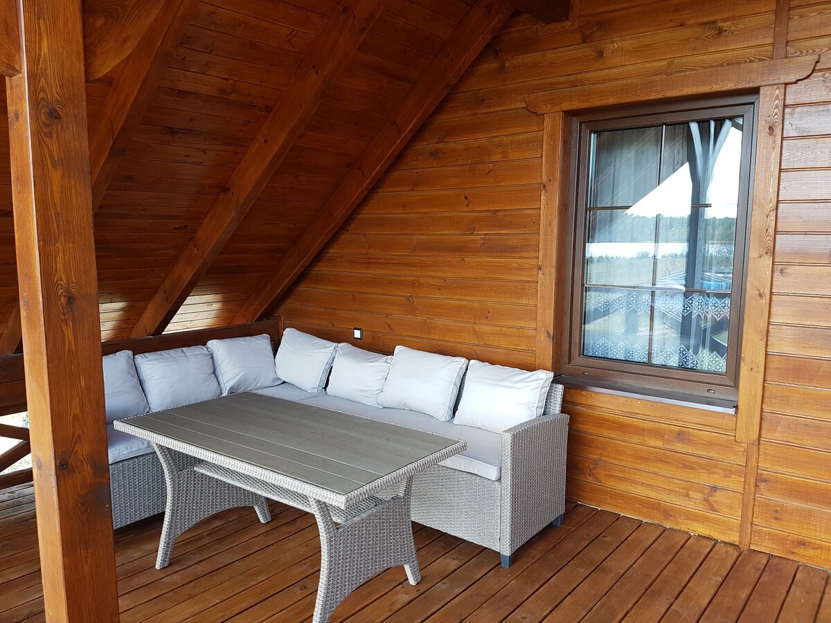 A terrace with a wooden floor features a comfortable seating area consisting of a light-colored sectional sofa adorned with plush cushions. A rectangular table stands in front of the seating, while a window with a decorative curtain frames the scenic outdoors.