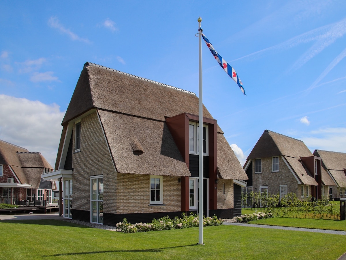 A charming villa is displayed, featuring a thatched roof and a well-maintained lawn. A flagpole adorned with colorful flags stands in front, while neighboring houses with similar architectural styles complete the scene under a bright blue sky.