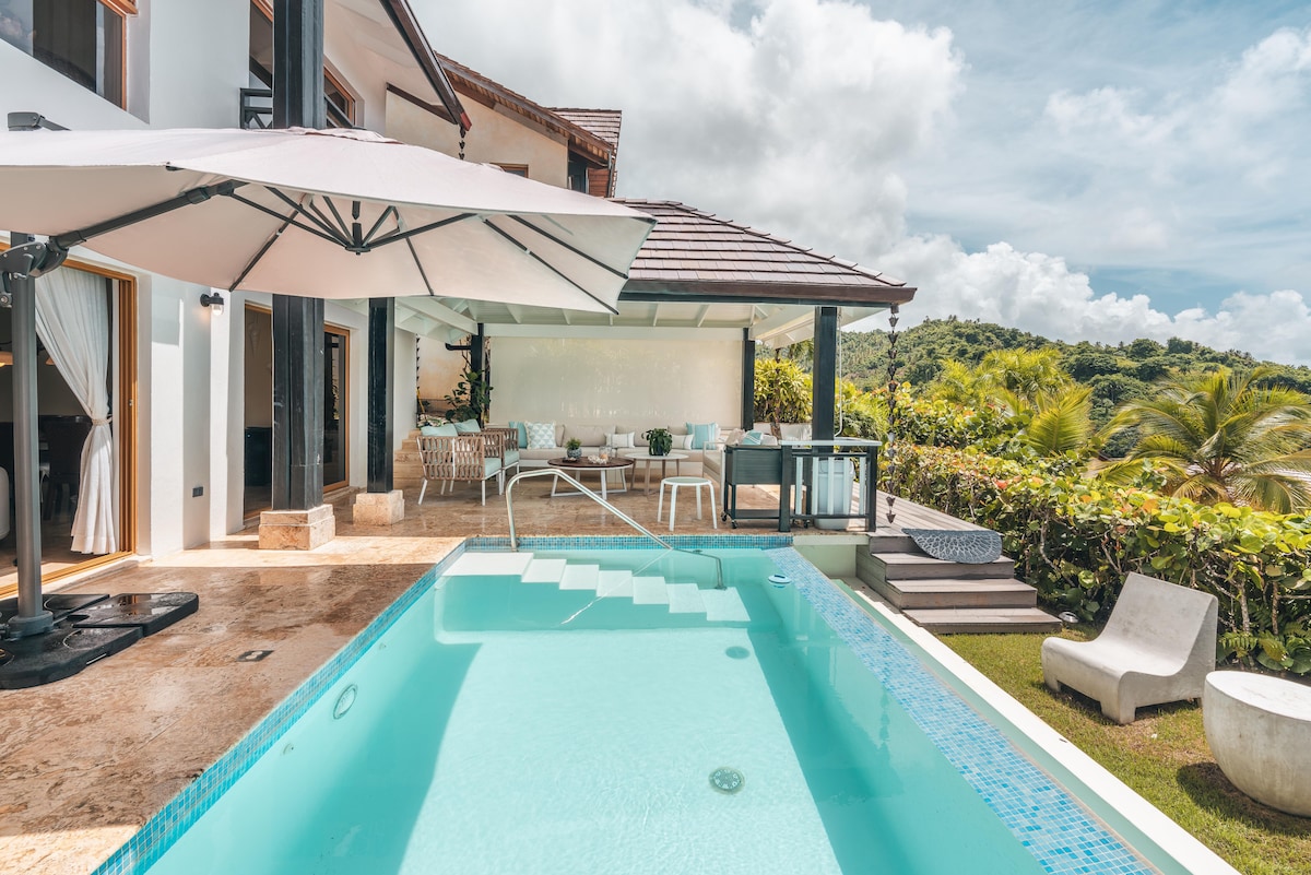 A private swimming pool is set against a backdrop of lush greenery, with steps leading into the crystal-clear water. Comfortable outdoor seating is arranged under a large umbrella, providing shade for relaxation. Nearby, a spacious terrace offers a serene view of the surrounding landscape.