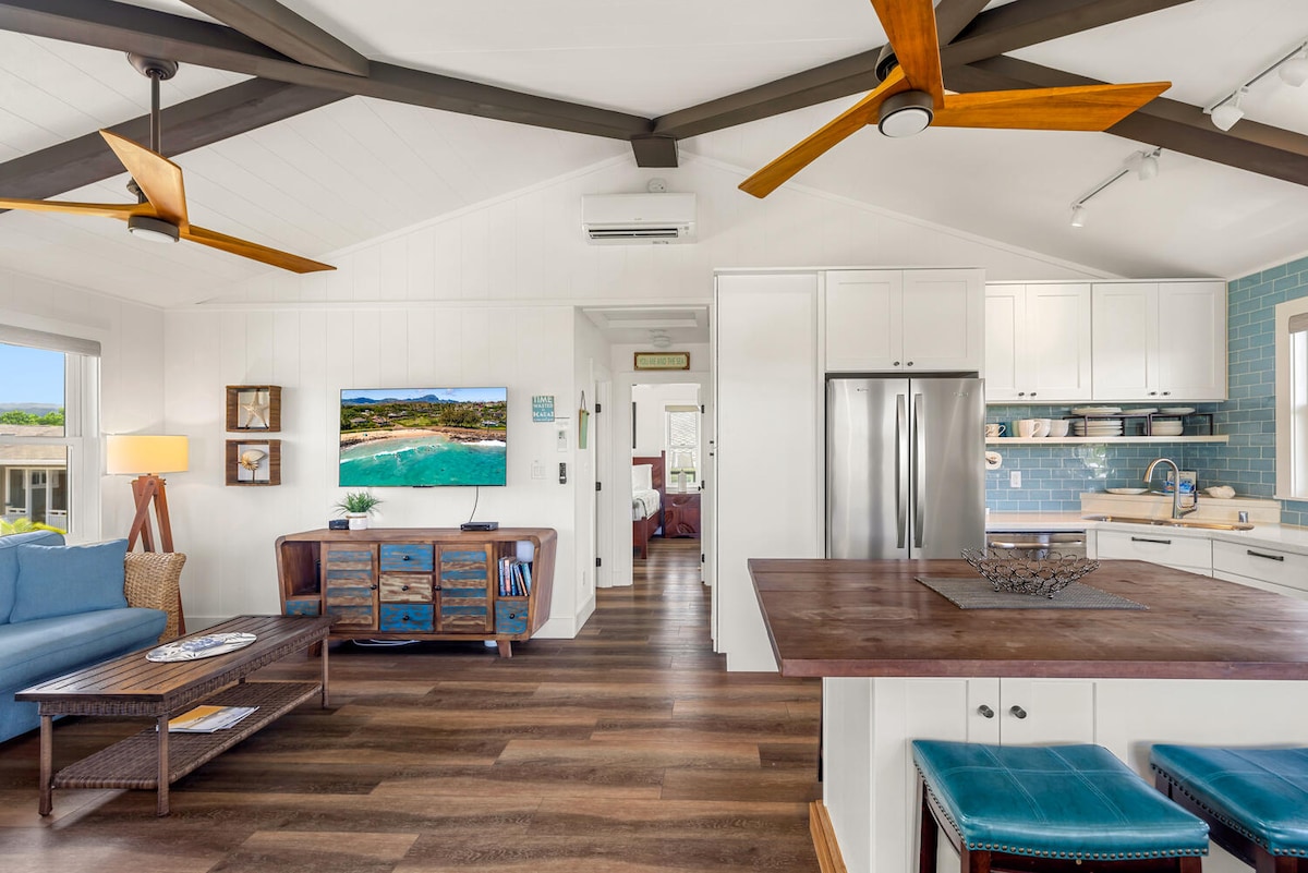 An open-concept living area features wood beams and light-colored walls, creating a bright atmosphere. A comfortable blue sofa faces a mounted television, complemented by a wooden coffee table. A kitchen with modern appliances is visible, along with a dining area set against a backdrop of sunny windows.