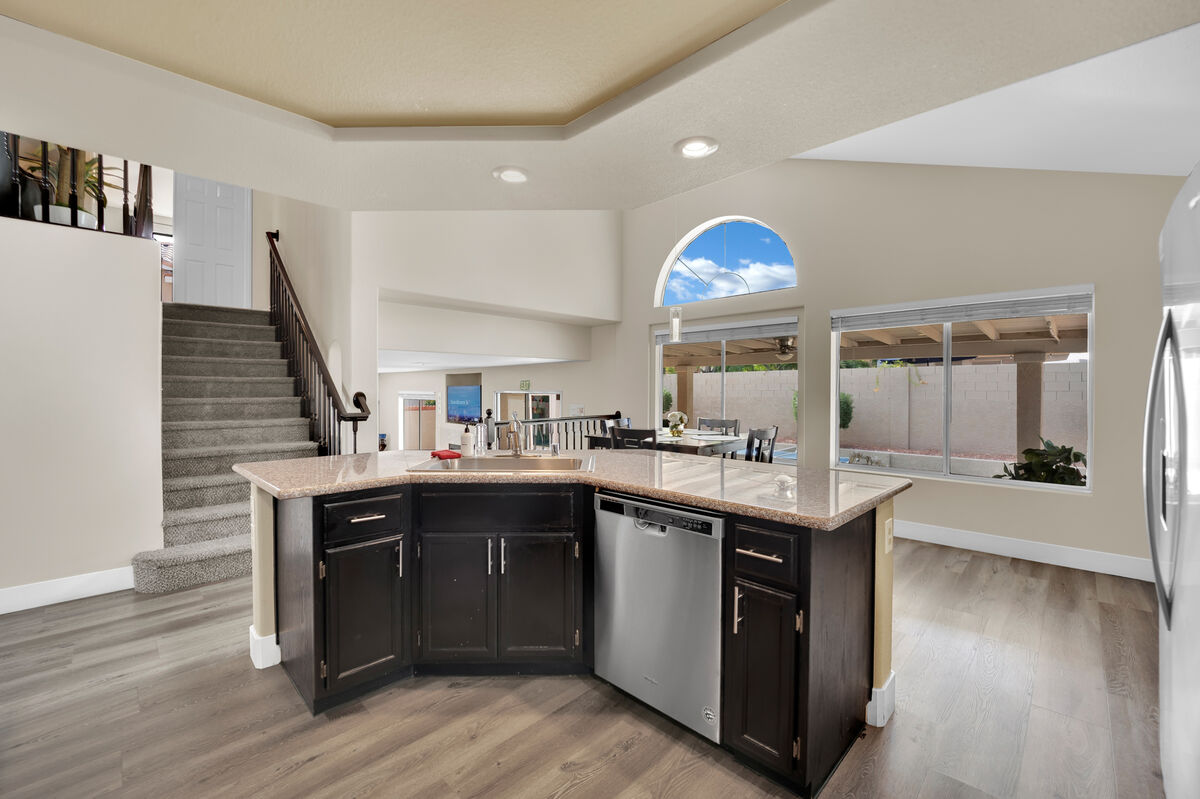 A modern kitchen space features an island with a granite countertop and stainless steel dishwasher. Large windows allow natural light to fill the area, creating an open and inviting atmosphere. The staircase and living area are visible in the background, enhancing the spacious layout.