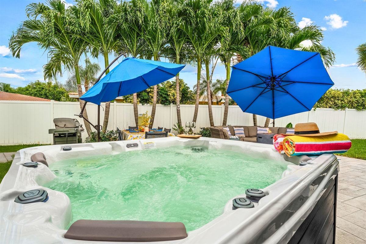 A hot tub is positioned in a spacious backyard, surrounded by lush palm trees. Two large blue umbrellas provide shade, while a colorful towel rests on the edge. The clear water invites relaxation in a tranquil outdoor setting.