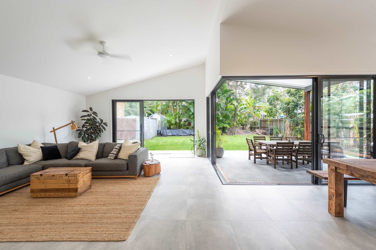 A modern living area features a spacious, comfortable lounge with a neutral color palette and a wooden coffee table. Large glass doors open to a well-maintained outdoor entertaining space, framed by lush greenery, creating a seamless connection between indoor and outdoor living.