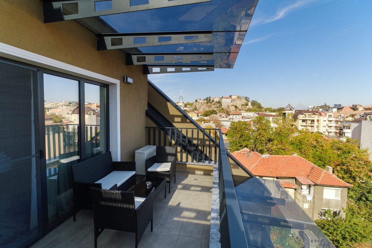 A balcony area features modern wicker furniture, including a table and two chairs. The space is covered by a glass awning, while spacious city views showcase nearby rooftops and greenery. The railing offers safety, and the area appears well-lit by natural sunlight.