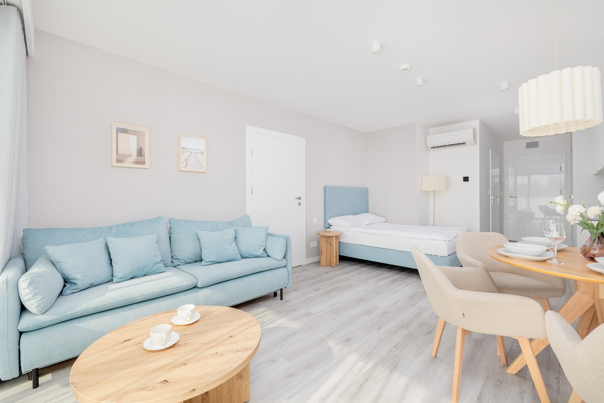 The well-designed studio features a light-colored interior with a blue sofa and a spacious double bed. A round wooden table with chairs is set for dining, while natural light fills the room through large windows. Air conditioning is positioned above the bed for comfort.