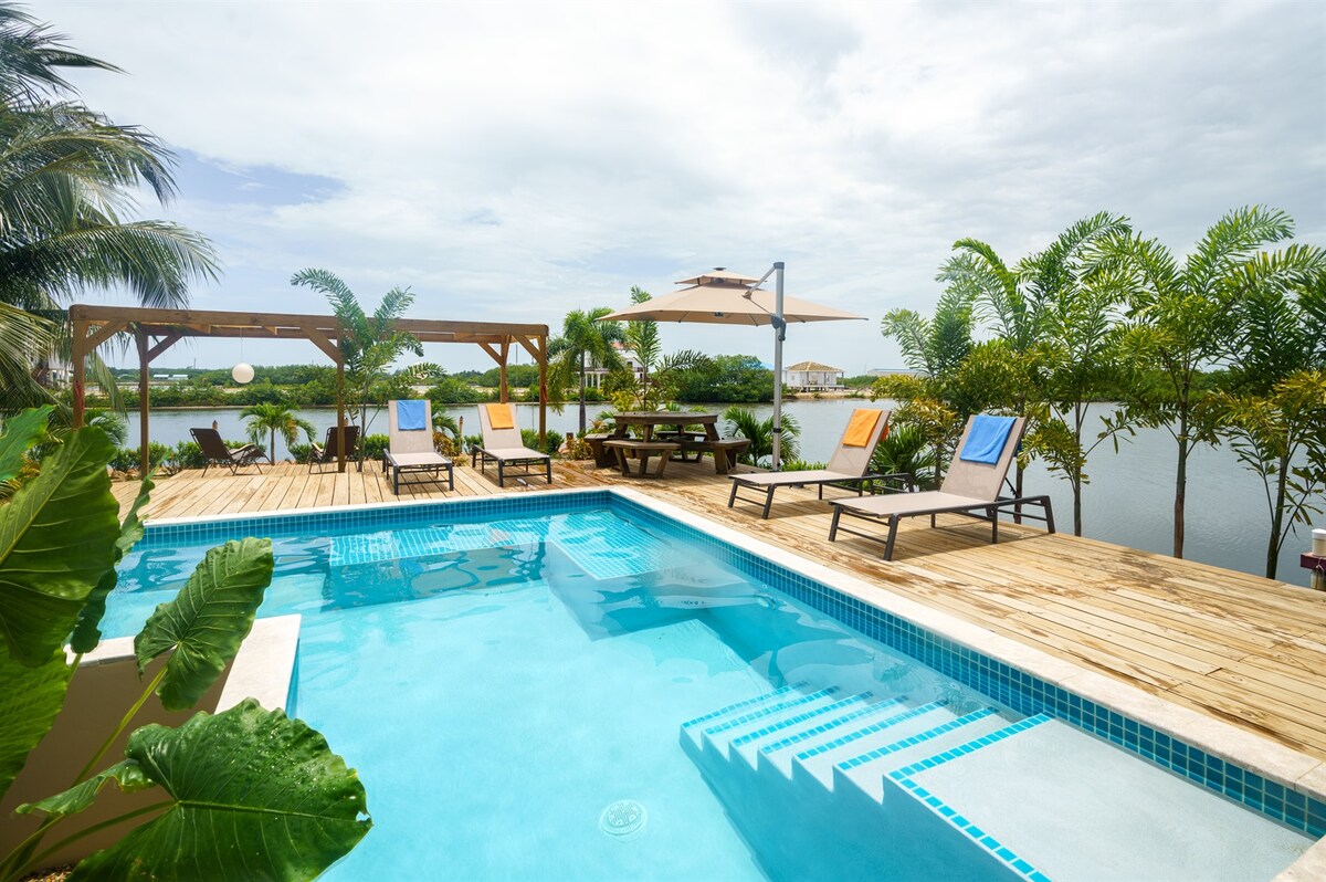 A serene outdoor pool area is framed by tropical greenery. Comfortable sunbeds are positioned on a spacious wooden deck, with an outdoor dining area sheltered by a pergola. The calm water of the bay is visible in the background, enhancing the tranquil atmosphere.