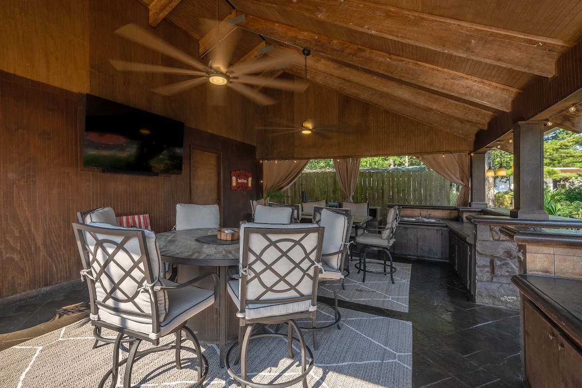The covered outdoor patio features a large circular table surrounded by six cushioned chairs. A bar area with additional seating is visible, along with a mounted TV. Soft lighting and ceiling fans enhance the comfortable atmosphere, creating an inviting space for gatherings.