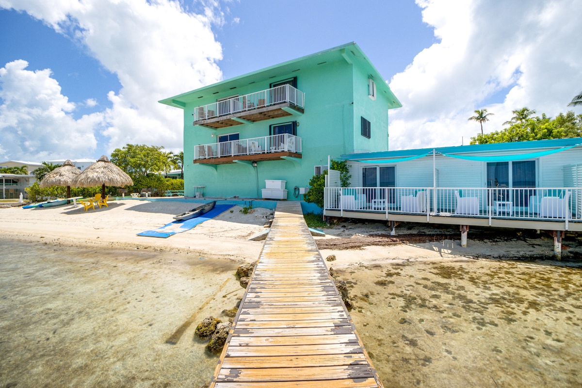 A light blue building is positioned near the water, with two stories and multiple balconies. A wooden boardwalk leads from the shore to the entrance, lined with lounge chairs and protected by thatched umbrellas. The clear water reflects the bright sky above.