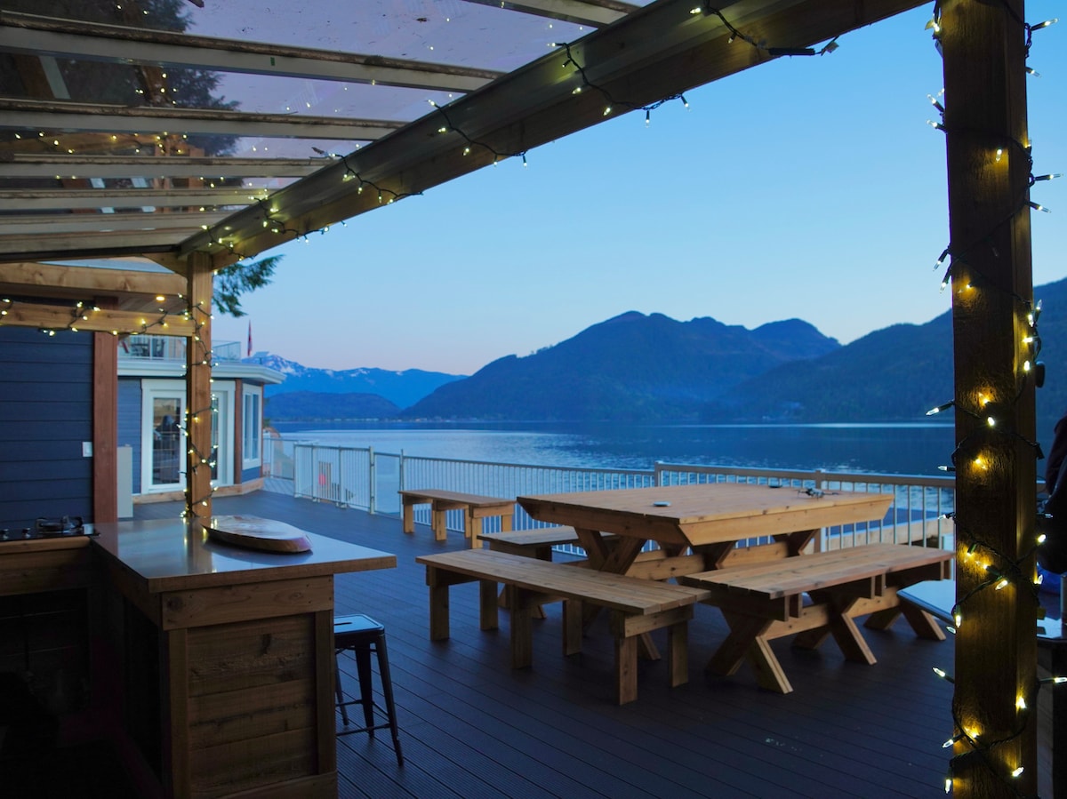 An outdoor seating area features large wooden picnic tables surrounded by a railing. String lights are visible along the structure, providing a warm ambiance. A serene view of the mountains and water is seen in the background under a clear blue sky.