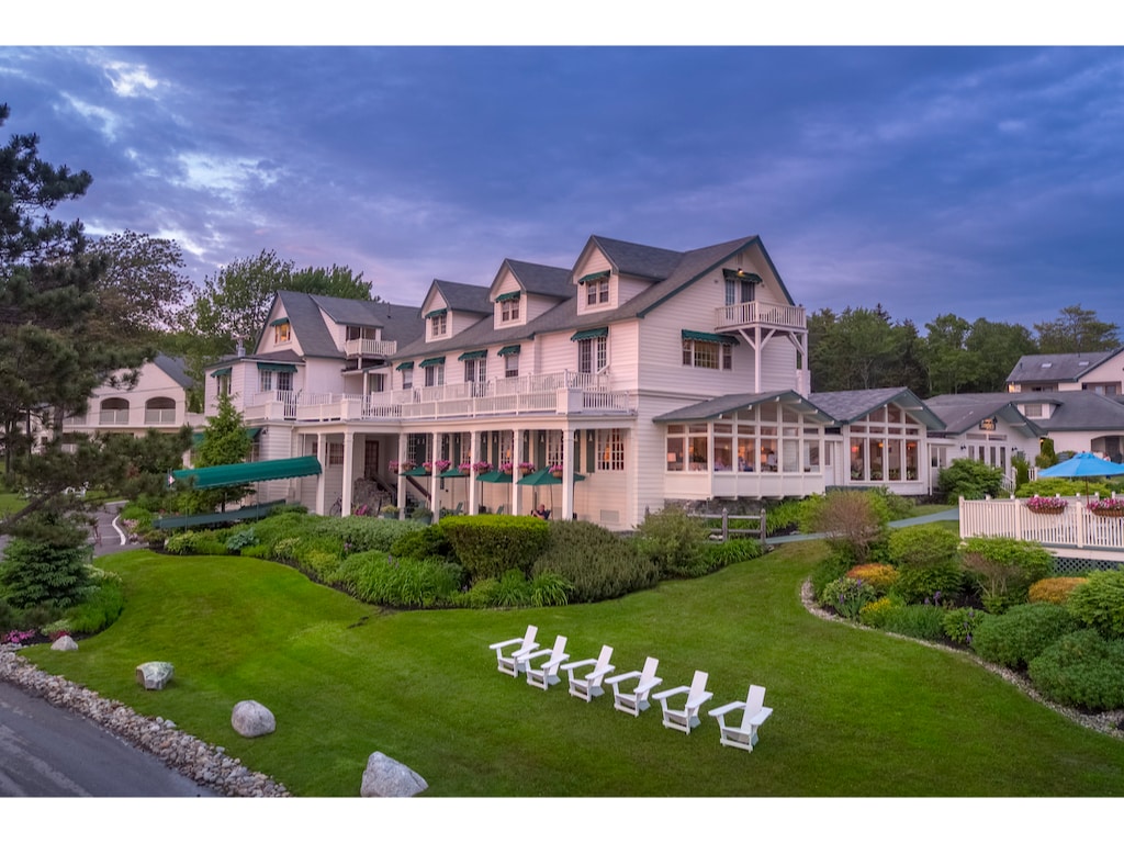 A multi-story building is set amid well-maintained gardens, with green lawns and pathways visible. The exterior features a light color scheme with multiple gabled roofs. Adirondack chairs are arranged on the lawn, inviting relaxation, while vibrant flower planters enhance the welcoming atmosphere.