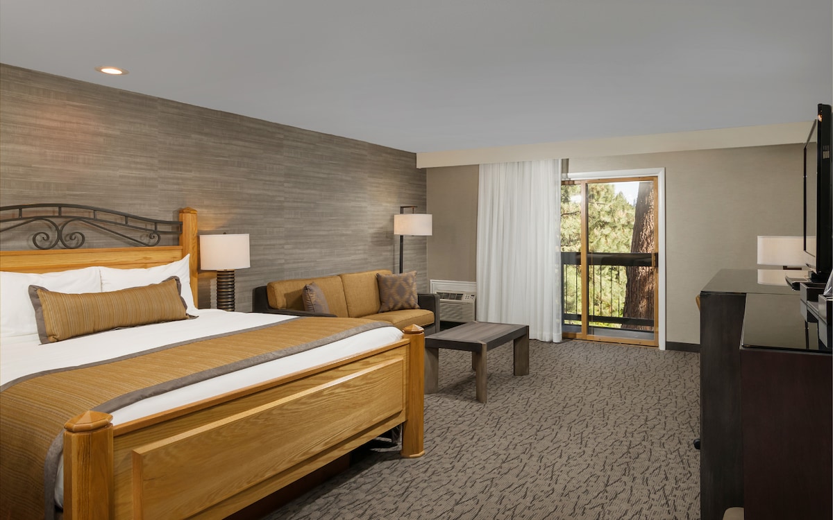 A spacious room is presented with a king bed dressed in warm-toned bedding. A sofa sits alongside a coffee table, complemented by bedside lamps. A large window offers a view of the outdoors, while a flat-screen television and a console unit complete the living space.