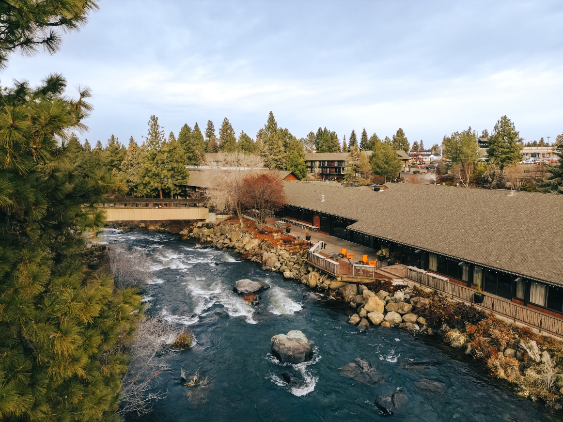 A scenic overview of the Riverhouse Lodge is presented, showcasing the lodge's wooden structure alongside the flowing Deschutes River. Trees surround the area, while rocky outcrops can be seen in the water. Outdoor seating is visible on the deck, inviting guests to enjoy the natural beauty.