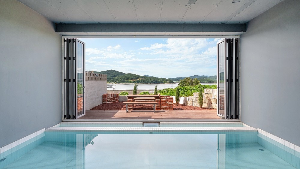 A serene indoor pool area features large folding glass doors that open to a spacious outdoor deck. Scenic views of the surrounding hills and water are visible, creating a seamless connection between the indoor and outdoor spaces. The pool reflects the natural light from the open doors.