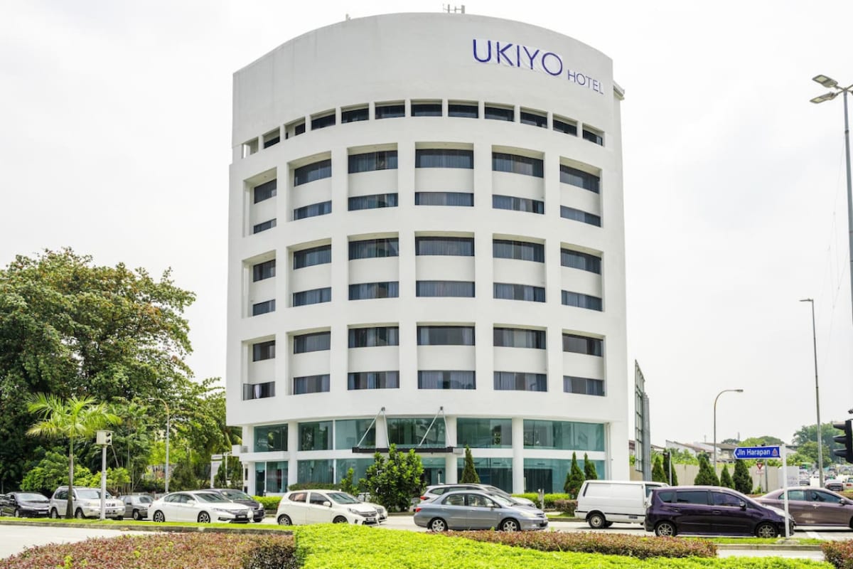 The entrance of Ukiyo Hotel is featured, showcasing a modern, six-story building with large windows. Surrounding greenery and a landscaped area enhance the welcoming appearance. Several parked cars are visible in the foreground, indicating accessibility and convenience in the hotel's location.