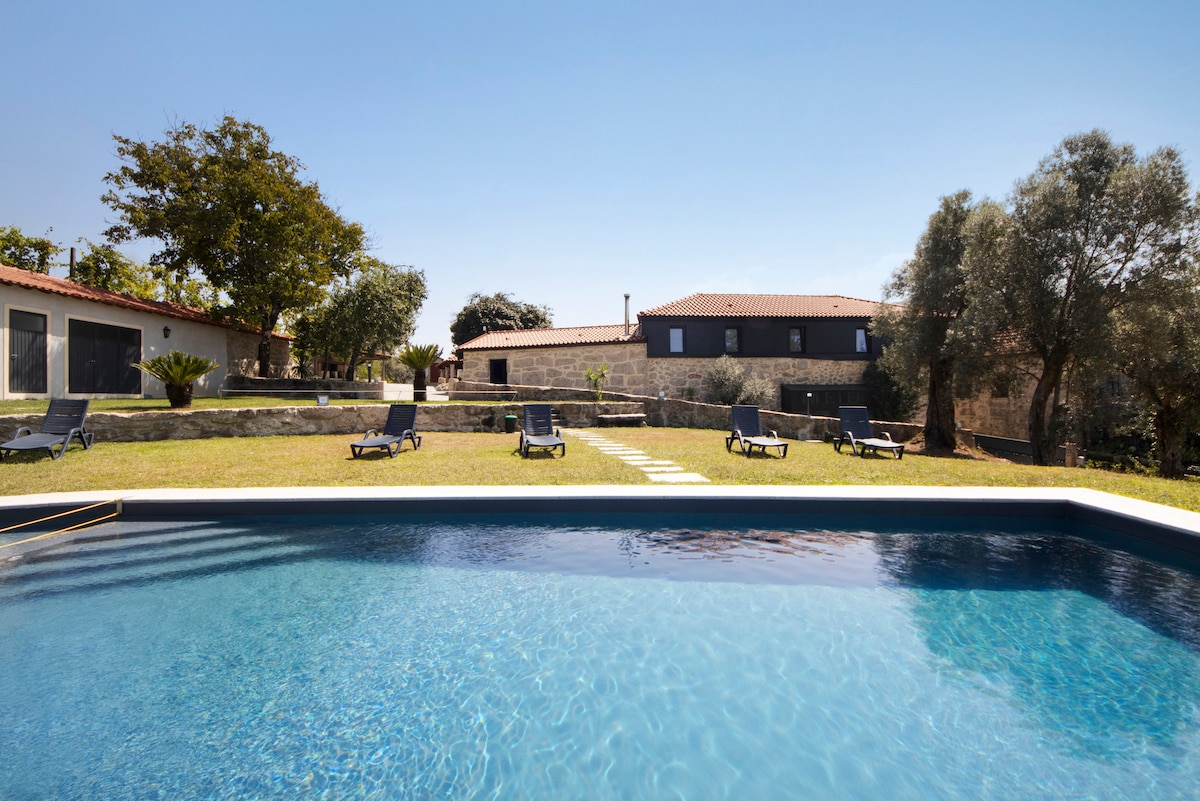 A private pool is surrounded by a grassy area, with sun loungers positioned for relaxation. In the background, the two-story country house and landscaped garden are visible, framed by mature trees under a clear blue sky.