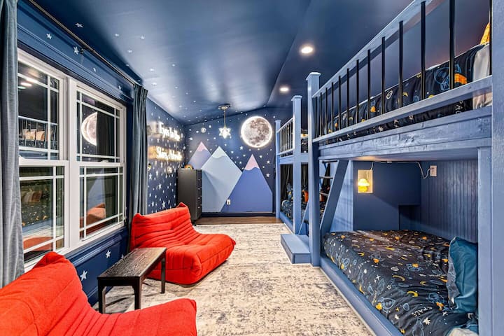 Esta habitación infantil con temática de astronauta cuenta con literas personalizadas con ropa de cama galáctica y una brillante luz de la pared de la luna, lo que crea una acogedora aventura espacial. Las paredes azules profundas de la habitación, los detalles estrellados y las vibrantes sillas rojas completan el ambiente celeste.