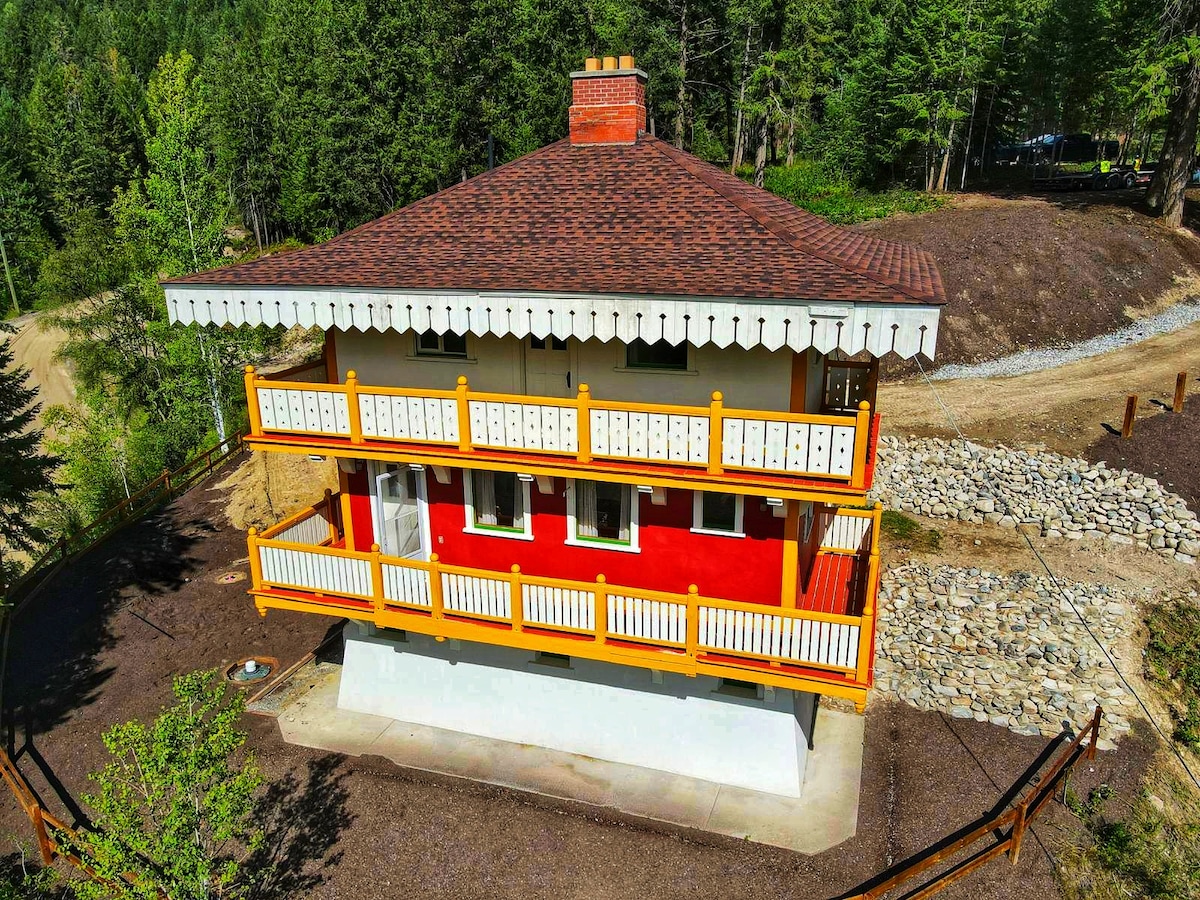 The exterior of the chalet is presented from an aerial perspective, highlighting its Swiss-inspired architecture. A vibrant red facade is complemented by white trim and an overhanging roof adorned with decorative brackets. Surrounding nature features green trees and gravel pathways, enhancing the tranquil setting.