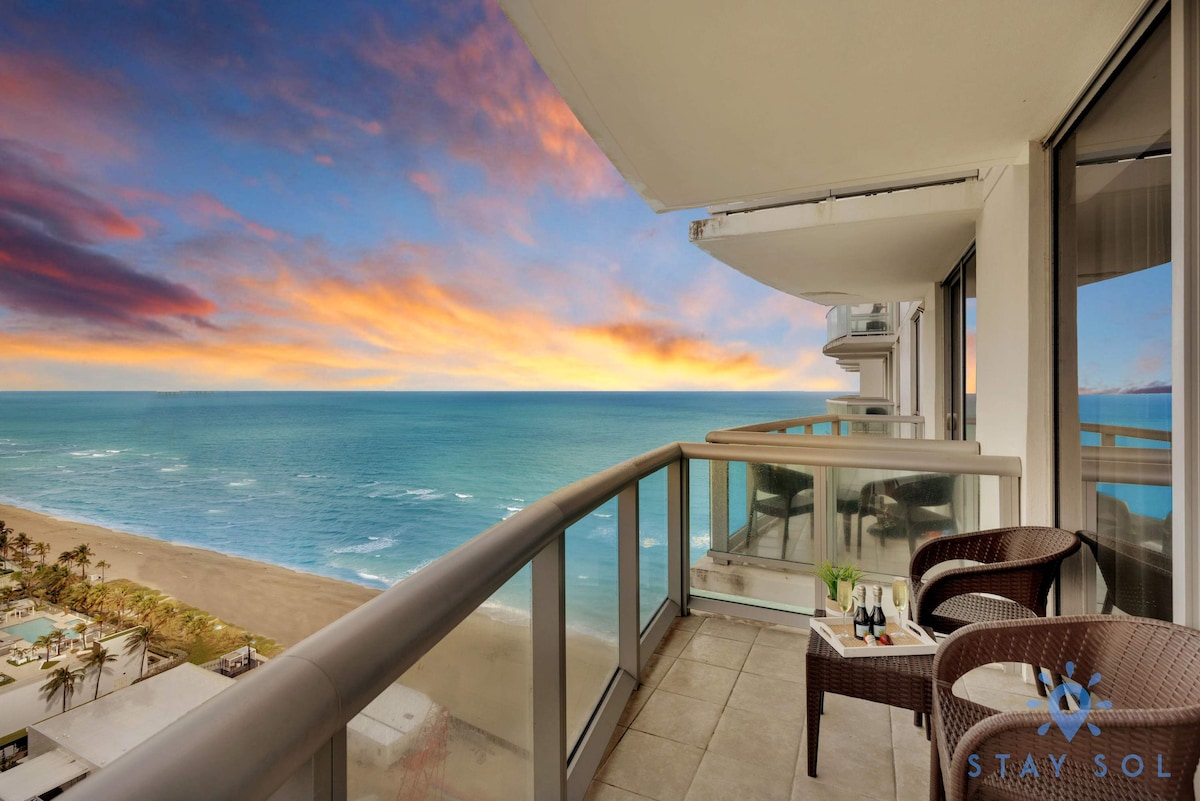 The balcony features two wicker chairs with a small table, overlooking an expansive view of the ocean and sandy beach. The vibrant sky at sunset casts a warm glow over the water, creating a peaceful outdoor space.
