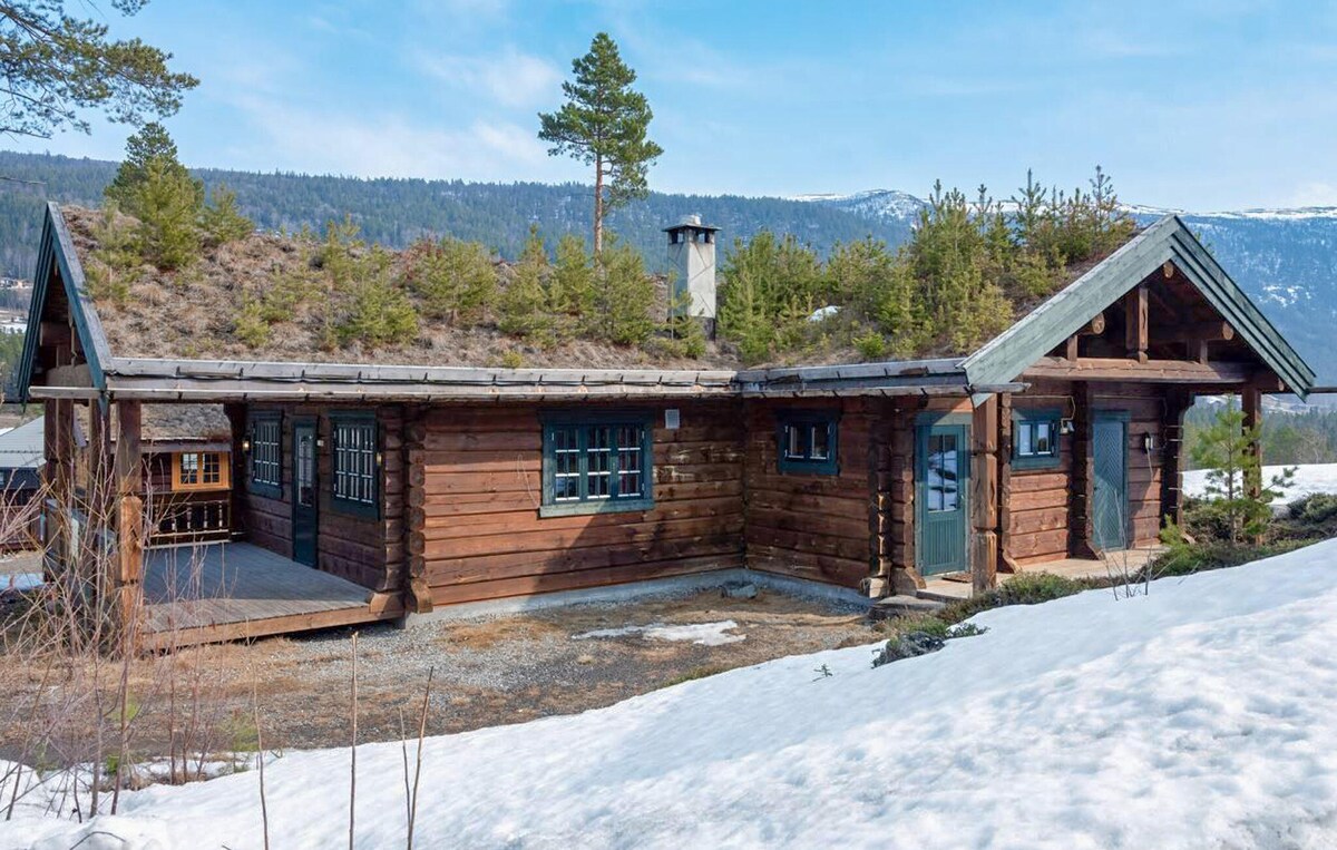 A wooden holiday home is surrounded by snow, showcasing a combination of natural materials and a sloped green roof adorned with small trees. The structure features large windows, a porch area, and a charming rustic design.