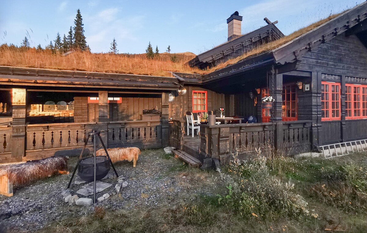 A charming wooden holiday home is featured with a traditional grass roof. A spacious terrace boasts red window frames, rustic furniture, and a fire pit surrounded by seating. The serene landscape includes grassy patches and trees in the background, creating a tranquil outdoor setting.