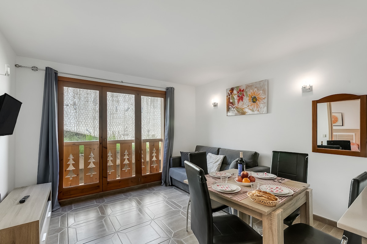 An open-plan living area is featured, with a modern dining table set for four and a comfortable sofa positioned nearby. Light enters through large windows adorned with decorative panels, illuminating the space. A flat-screen TV and a wall mirror enhance the ambiance.