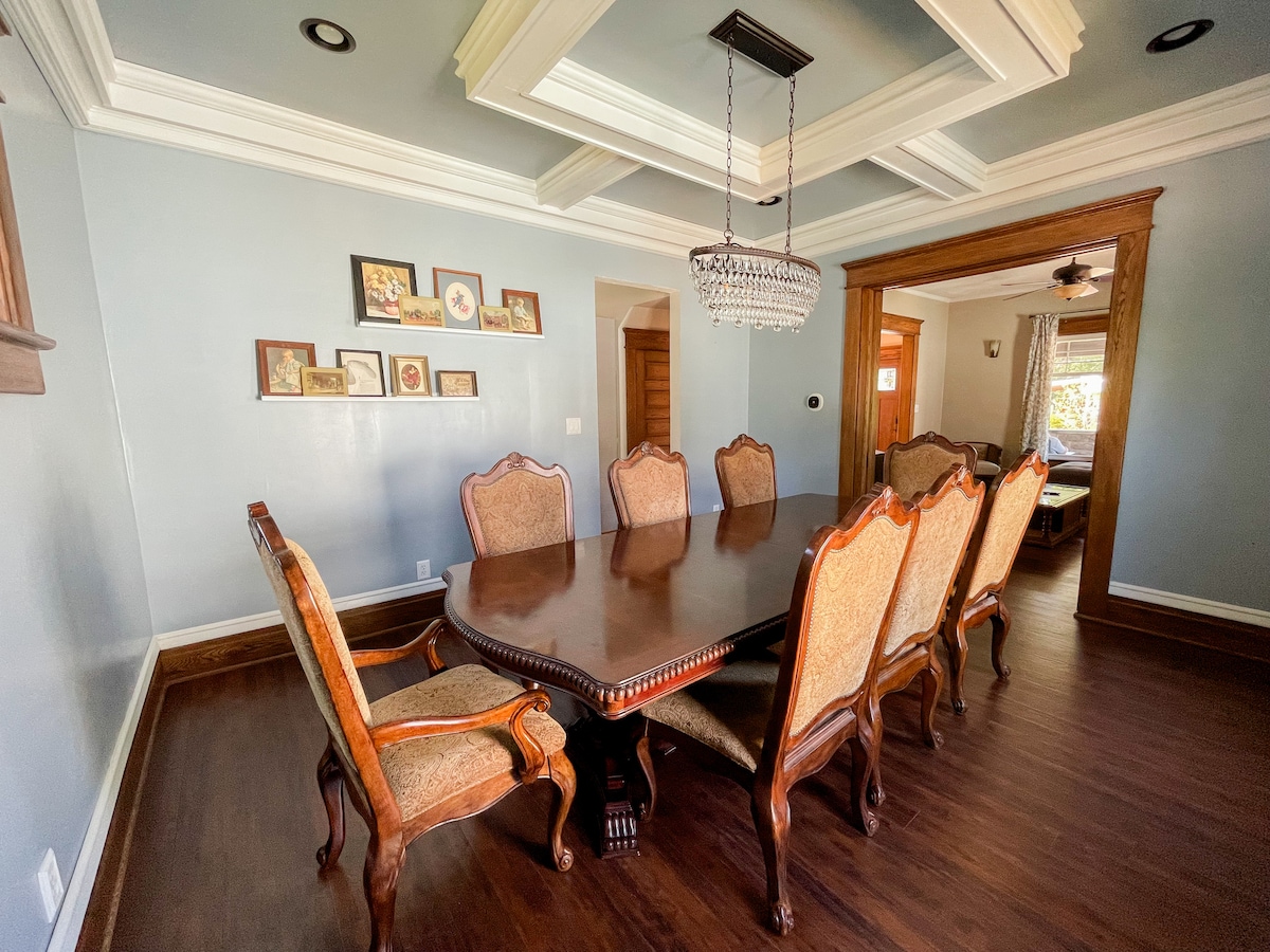 A large dining room is presented, featuring a meticulously crafted wooden dining table surrounded by eight intricately designed chairs. A chandelier hangs elegantly from the ceiling, while artwork adorns the wall, adding character to the space.