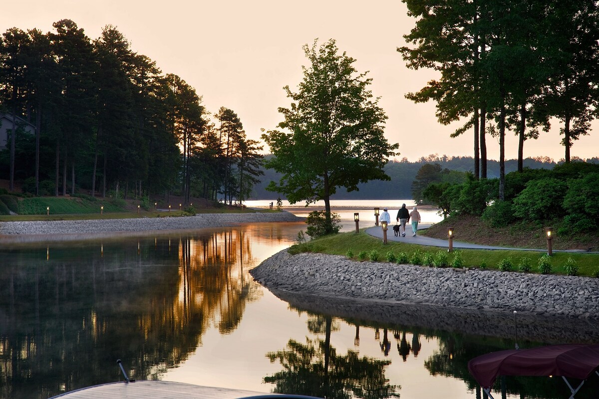 A serene lakeside path is lined with trees, reflecting on still water as the sun sets. Soft lighting from pathway lamps casts a gentle glow along the edge, where a couple walks with their dog. The tranquil scene is enveloped in the colors of dusk.