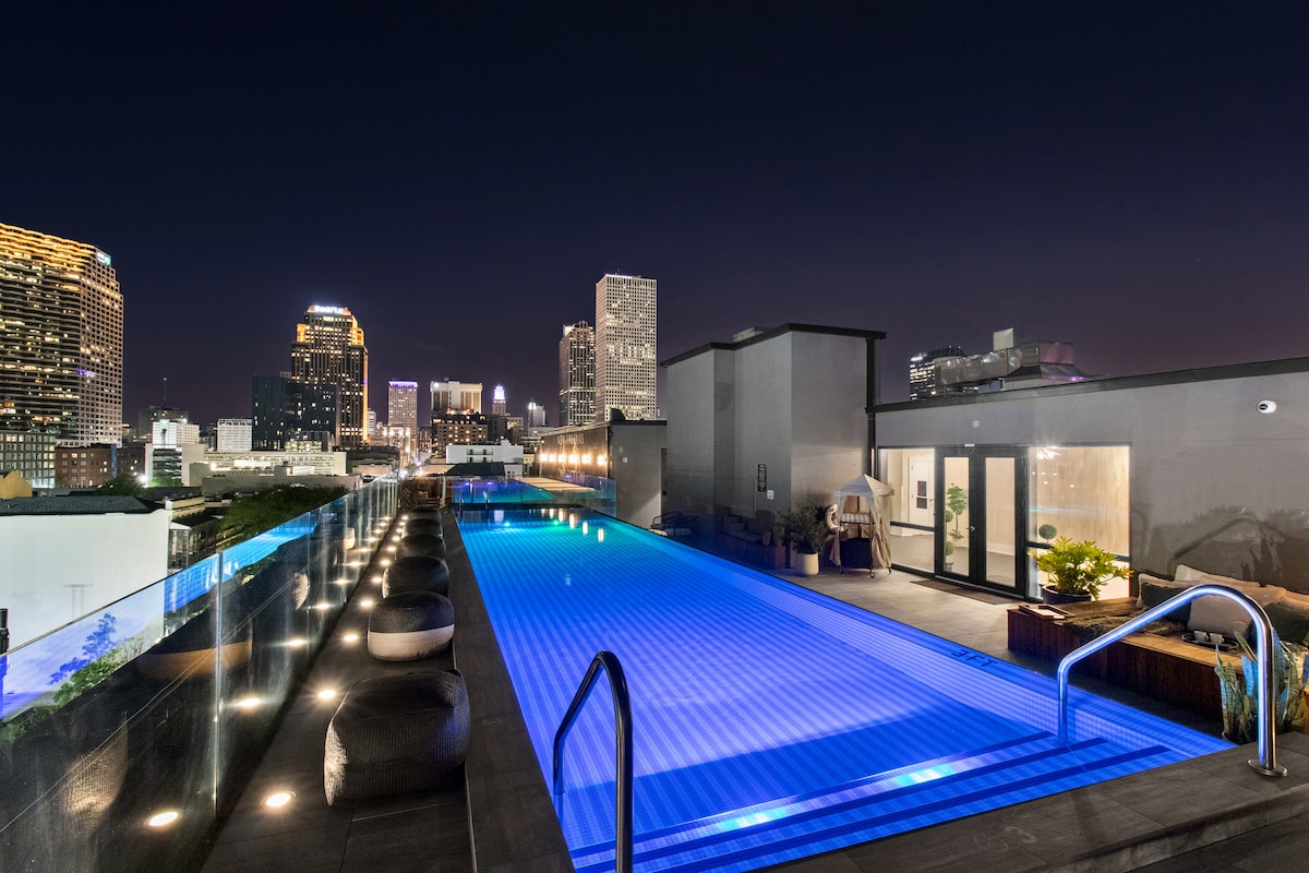 An illuminated rooftop pool features modern blue lighting that contrasts with the city skyline at night. Seating is available along the edge, providing a relaxing space with views of the surrounding buildings and vibrant urban landscape.