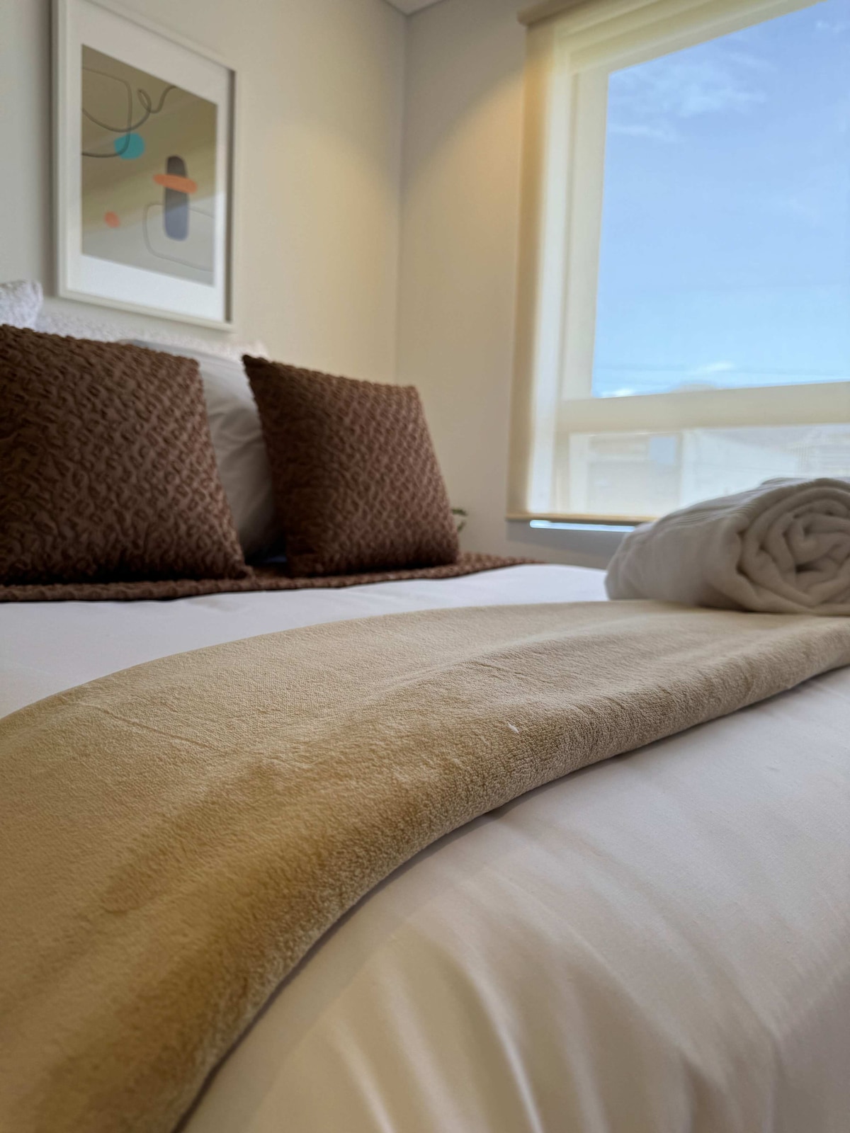 A spacious bed is adorned with two textured brown pillows and a light beige blanket neatly draped across it. A rolled white towel rests beside the pillows, and natural light streams in through a partially open window, illuminating the relaxing space.