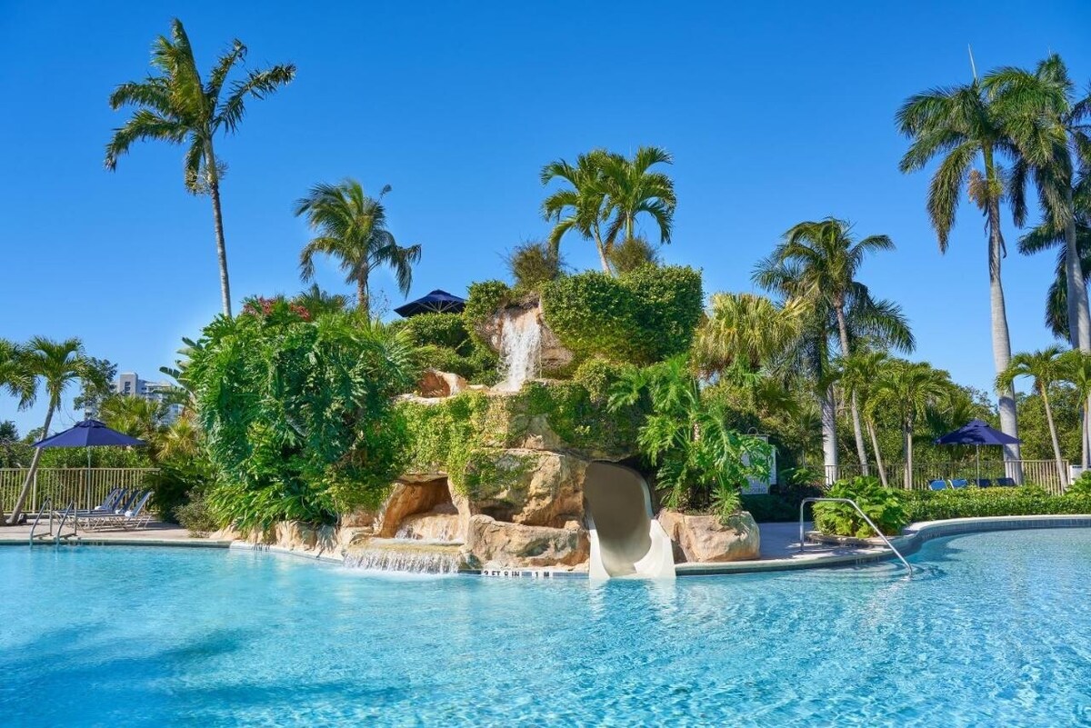 A large outdoor pool is surrounded by palm trees and lush landscaping, featuring a natural rock formation with a cascading waterfall and a waterslide. Sun loungers are positioned nearby, creating a relaxing atmosphere under the bright blue sky.