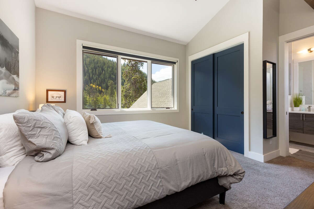 Un dormitorio con una cama grande, puertas azules en el armario y una ventana que muestra una vista de los árboles y una montaña. La habitación es luminosa con una decoración neutra.