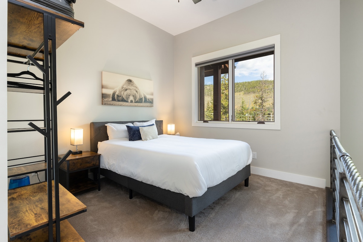 Dormitorio con una cama cuidadosamente hecha, sábanas blancas, dos mesitas de noche de madera con lámparas, un estante de metal y un gran ventanal con vista a los árboles y una montaña.