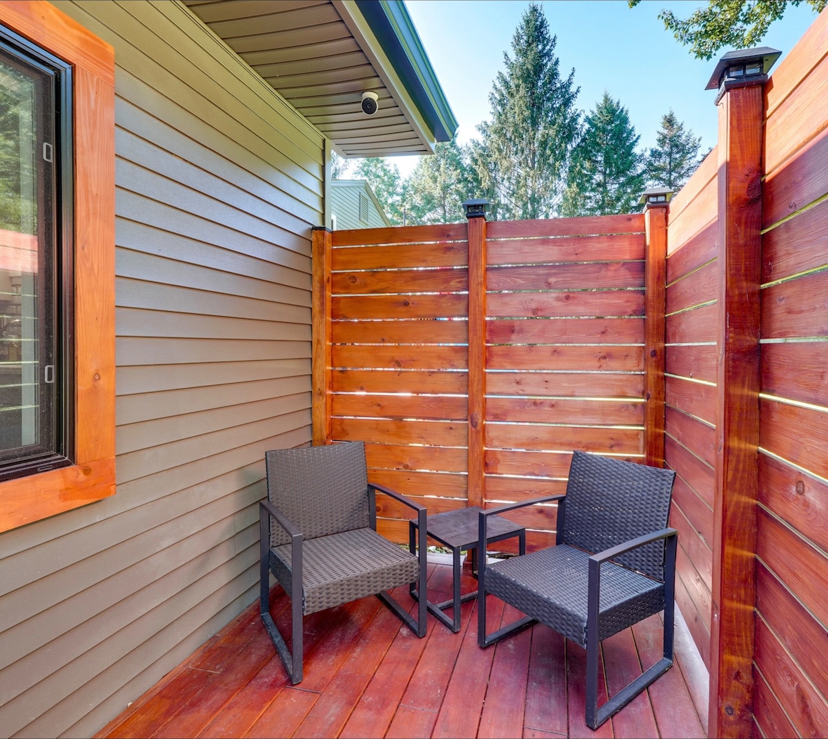 A private outdoor seating area features two dark wicker chairs and a small table, surrounded by wooden fencing for added privacy. Sunlight filters in, highlighting the wooden deck and the greenery in the background.