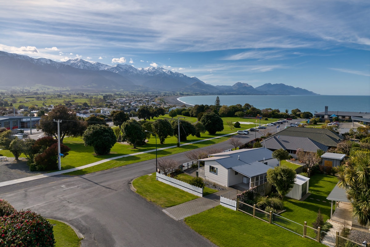 A panoramic view of the Kaikōura coastline, showcasing a mix of residential areas and natural landscapes. Snow-capped mountains rise in the background, while the ocean extends into the distance. Lush green lawns and trees line the roads, providing a serene atmosphere.