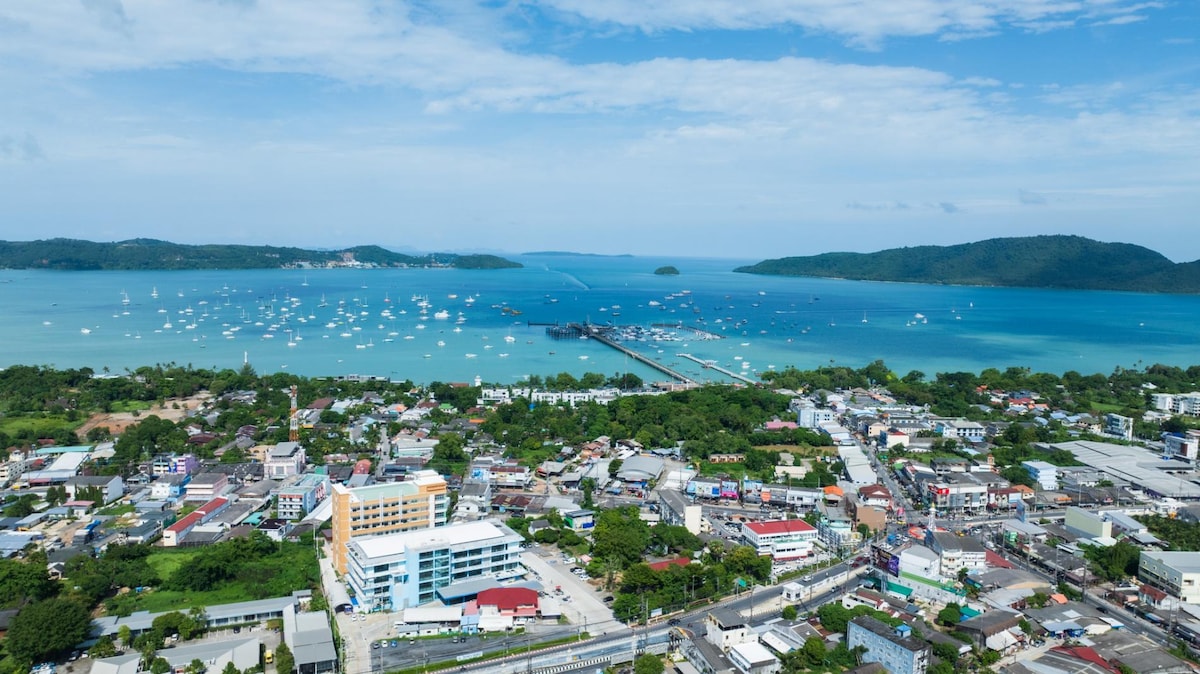 An expansive aerial view captures a vibrant coastal village bordered by the sea. Clear blue waters reflect the sky, dotted with boats. The shoreline showcases a mix of urban development and greenery, contributing to a lively atmosphere with residential and commercial buildings visible.