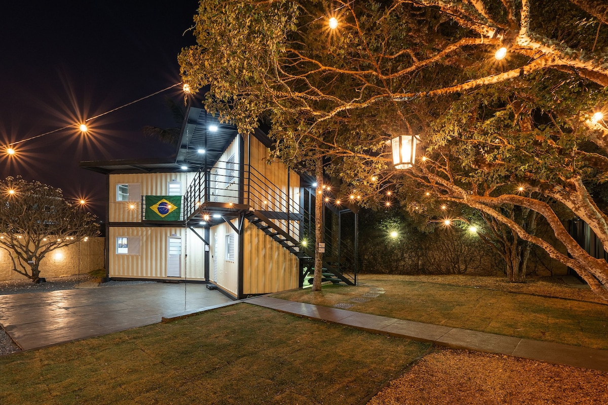 A container-style building is shown at night, illuminated by warm lights. The exterior features a Brazilian flag and two levels, with a staircase leading to the upper entrance. Surrounding greenery and a well-manicured lawn enhance the inviting appearance.