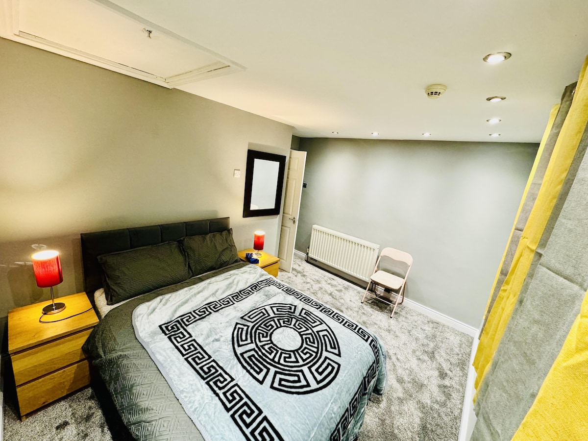 A well-lit bedroom features a spacious bed covered with a patterned blanket. Two bedside tables with lamps flank the bed. A neutral wall is complemented by a large mirror, and a chair is positioned by the wall. Soft carpeting adds comfort to the space.