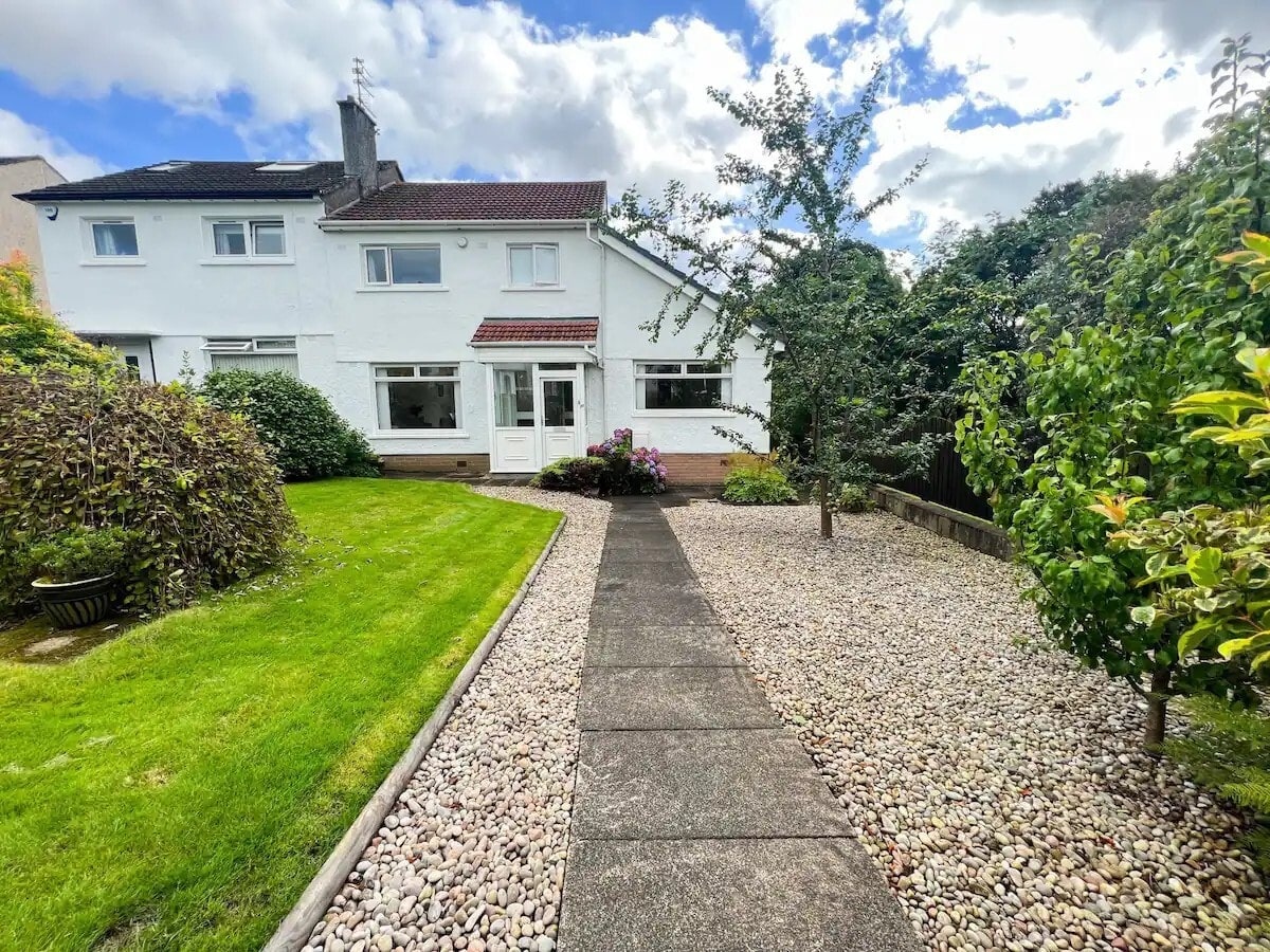 A well-maintained front garden leads to a private entrance of the residence. A pathway bordered by stone pebbles is flanked by lush greenery, flowering plants, and a tree, enhancing the welcoming atmosphere of the home's exterior.