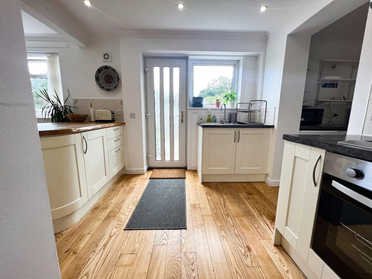 The bright open-plan kitchen features a stylish design with cream cabinetry and wooden flooring. A doorway with glass panels leads to the outdoor area, while modern appliances and ample counter space provide a functional cooking environment.