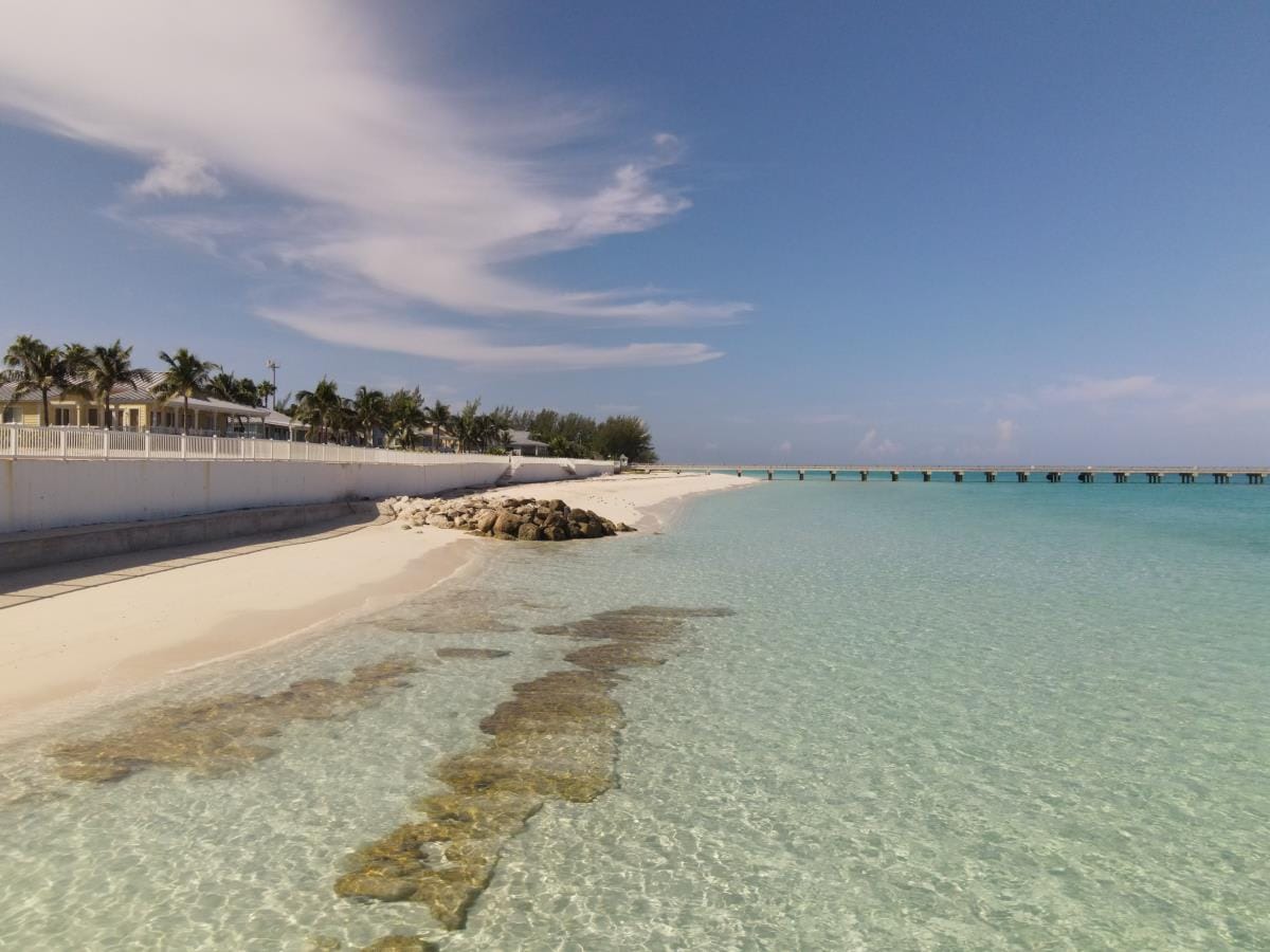 A serene beach scene is displayed with soft white sand and clear turquoise water. Gentle waves lap at the shoreline, revealing rocky formations beneath the surface. In the distance, a pier stretches out over the water, framed by palm trees that line the coast.