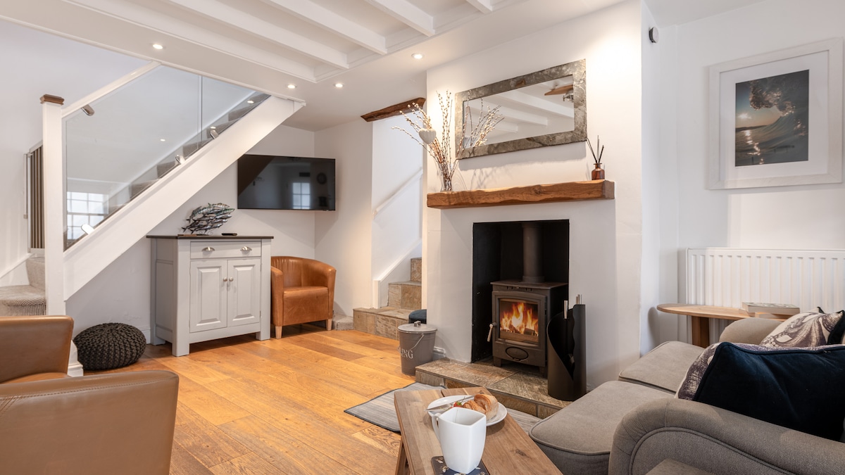 A welcoming ground floor living space features two two-seater sofas positioned around a coffee table. A wood burner sits beneath a rustic wooden mantle, complemented by a TV mounted on the wall. Step diverting to the kitchen area is visible to one side.