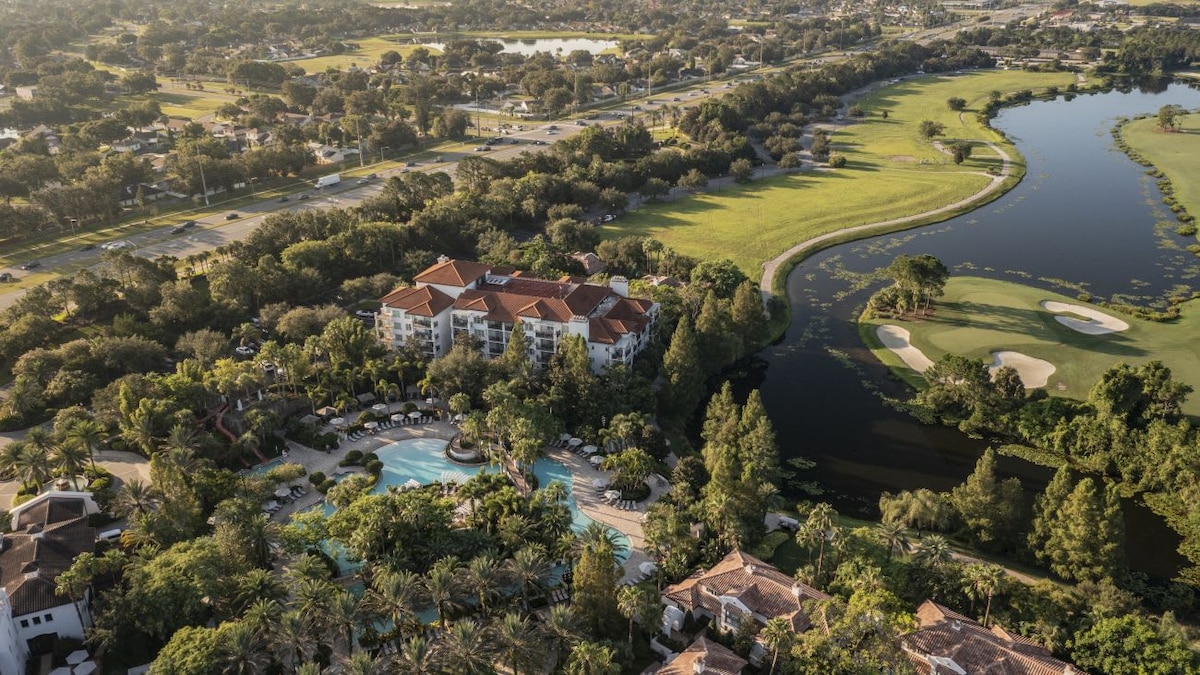An aerial view captures the resort amidst lush green landscapes, highlighting the swimming pool area surrounded by palm trees. Nearby, a golf course is visible, along with winding pathways leading towards a tranquil body of water. The building features a red-tiled roof consistent with the resort's architecture.