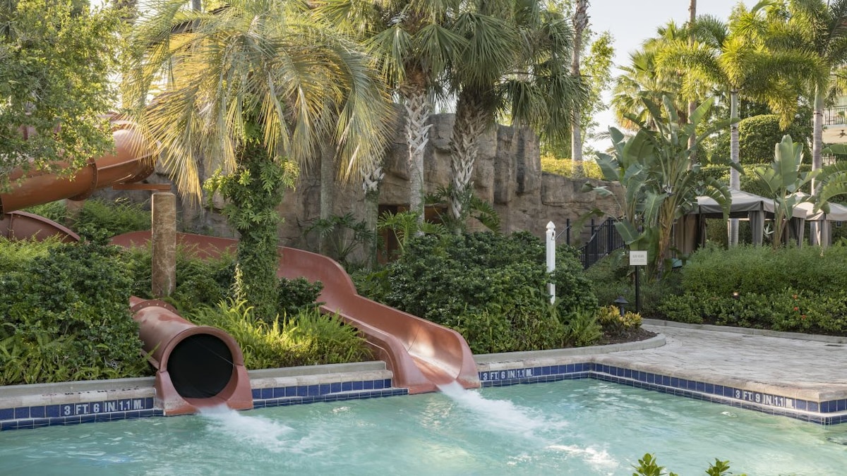 A water slide curves down into a clear swimming pool, surrounded by lush palm trees and tropical plants. The pool features a gently flowing water entrance, creating a fun and inviting atmosphere for guests of all ages.