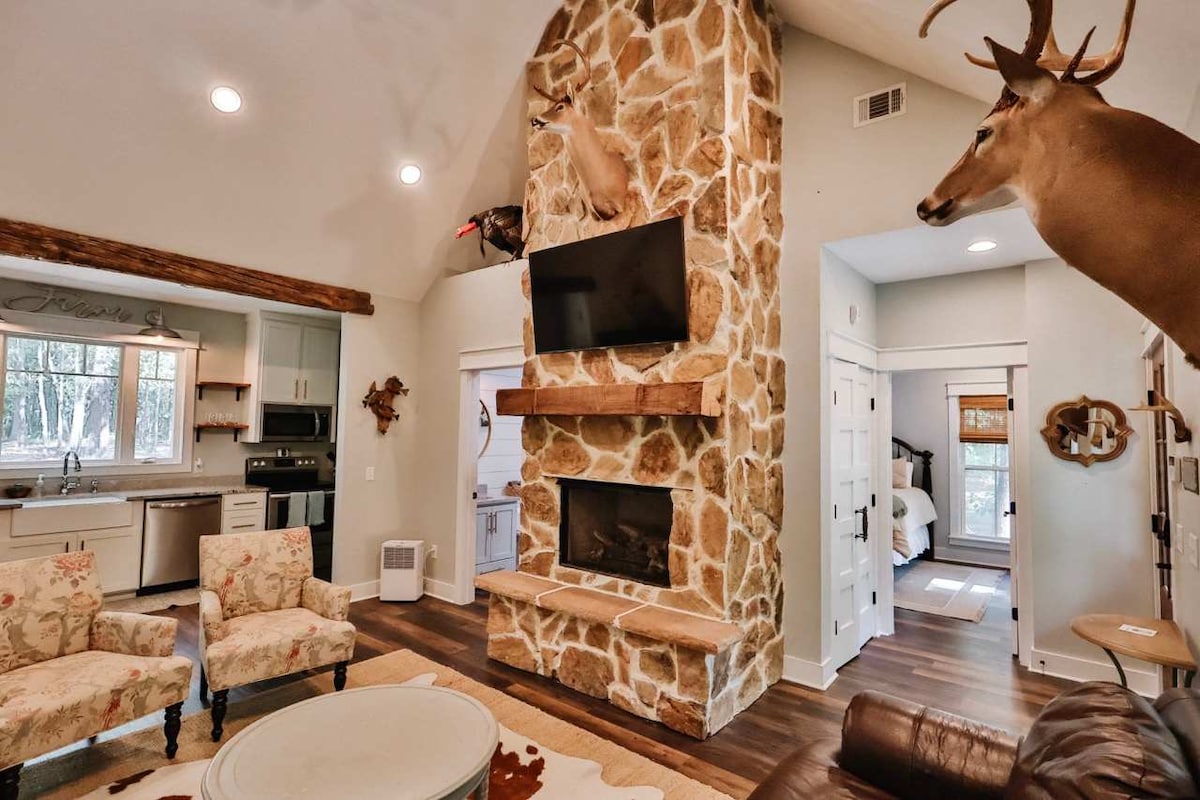 The living room showcases a stone fireplace with a mounted deer head above a wall-mounted TV. Two floral armchairs are positioned around a circular coffee table, complementing the rustic decor. A glimpse of the kitchen can be seen in the background, with modern appliances and natural light.