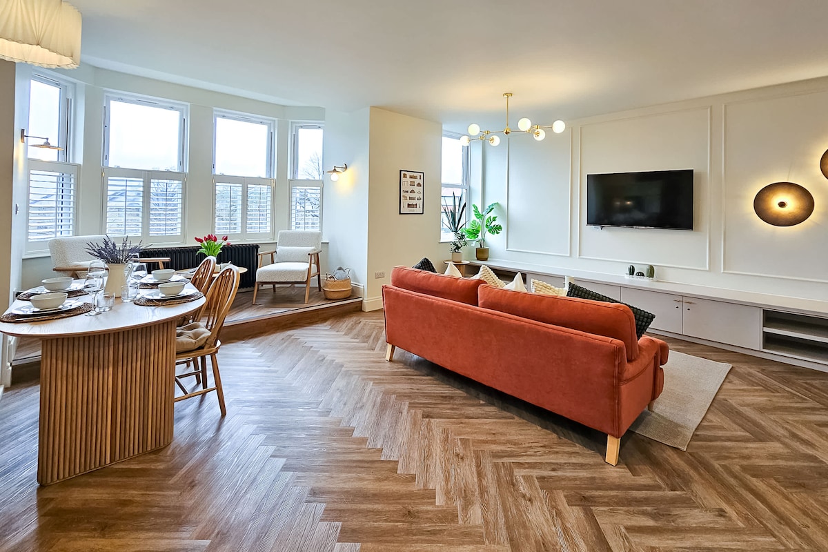 The open-plan living area features a warm-toned sofa and stylish furnishings. A flat-screen TV is mounted on the wall, and large windows allow natural light to brighten the space. A dining table is set for four, with modern decor and greenery enhancing the ambiance.